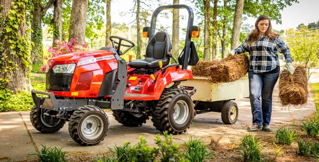 Massey Ferguson GC1700 Series sub-compact tractor hauling straw with a woman working beside it in a wooded residential setting
